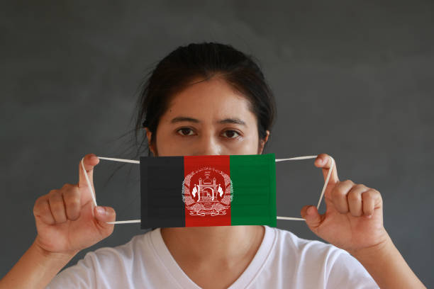 una donna in camicia bianca con bandiera afghana su maschera igienica in mano e sollevò la faccia anteriore su sfondo grigio scuro. - arms lifted foto e immagini stock