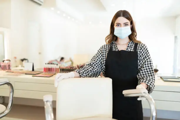 Female makeup artist wearing face mask while standing by chair in beauty salon