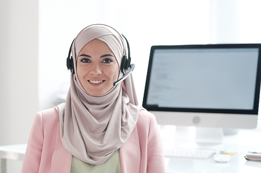 Portrait of positive beautiful young Muslim woman in hijab answering questions using hands-free device in call center
