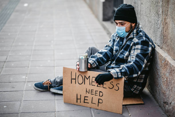 Homeless man sitting and begging for help Homeless young man sitting on sidewalk and begging alone. He is wearing protective face mask and holding out a can and begging for money. His head is bowed in shame and tiredness and hunger. homelessness photos stock pictures, royalty-free photos & images