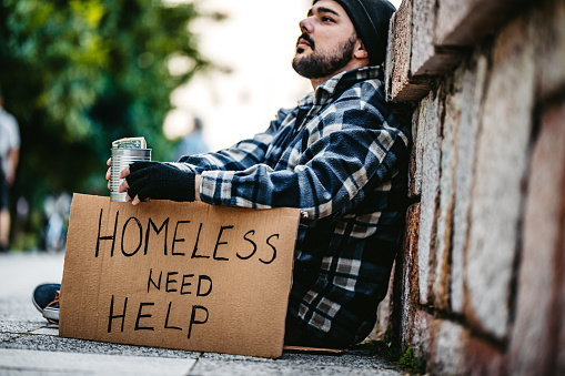 Homeless young man sitting and begging alone. He is holding out a can and begging for money. His head is bowed in shame and tiredness and hunger.