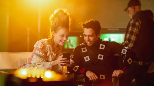 Photo of Beautiful Smiling Actress Wearing Renaissance Dress and Actor Wearing Motion Capture Suit Sitting on Chairs Share Social Media Posts via Smartphone. On Film Studio Period Costume Drama Film Set