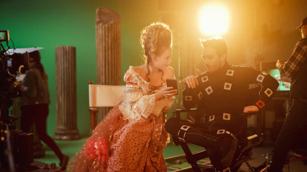 Beautiful Smiling Actress Wearing Renaissance Dress and Actor Wearing Motion Capture Suit Sitting on Chairs Share Social Media Posts via Smartphone. On Film Studio Period Costume Drama Film Set hermosa actriz sonriente que lleva vestido renacentista y actor que lleva traje de captura de movimiento sentado en las sillas comparten publicaciones en las redes sociales a través de smartphone. en film studio period traje drama película set