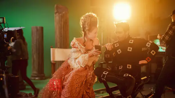 Photo of Beautiful Smiling Actress Wearing Renaissance Dress and Actor Wearing Motion Capture Suit Sitting on Chairs Share Social Media Posts via Smartphone. On Film Studio Period Costume Drama Film Set