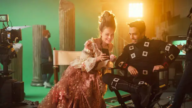 Photo of Beautiful Smiling Actress Wearing Renaissance Dress and Actor Wearing Motion Capture Suit Sitting on Chairs Share Social Media Posts via Smartphone. On Film Studio Period Costume Drama Film Set