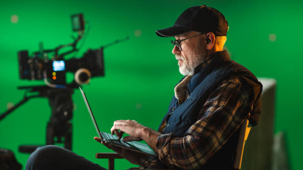 istock Prominent Successful Director Sitting in a Chair on a Break and Using Laptop Computer. On the Studio Film Set with High-End Equipment Professional Crew Shooting High Budget Movie 1265157400