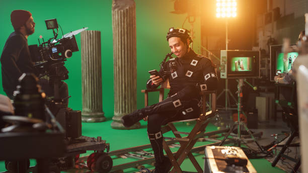 istock Handsome Smiling Actor Wearing Motion Capture Suit and Head Rig having Lunch Break, Sitting on Chair, Uses Smartphone. Studio High Budget Movie. On Film Studio Period Costume Drama Film Set 1265157441