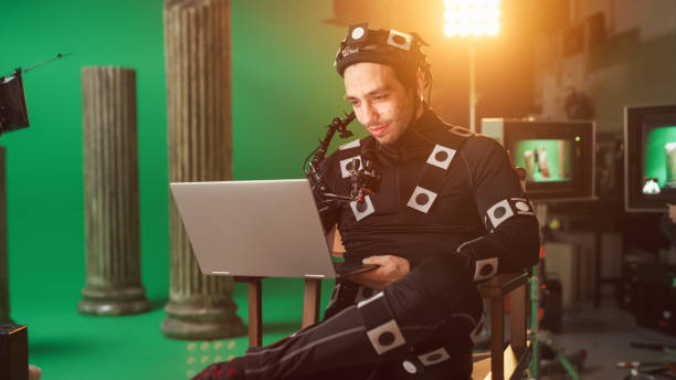 istock Handsome Smiling Actor Wearing Motion Capture Suit and Head Rig having Lunch Break, Sitting on Chair, Use Laptop. Studio High Budget Movie. On Film Studio Period Costume Drama Film Set 1265157444