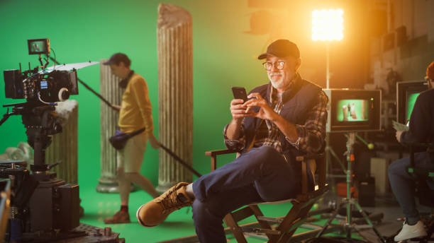 istock Prominent Successful Director Sitting in a Chair on a Break Using Smartphone. On the Studio Film Set with High-End Equipment Professional Crew Shooting High Budget Movie. 1265157454