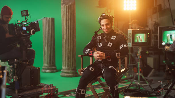 istock Handsome Smiling Actor Wearing Motion Capture Suit and Head Rig having Lunch Break, Sitting on Chair, Looking at Camera. Studio High Budget Movie. On Film Studio Period Costume Drama Film Set 1265157479