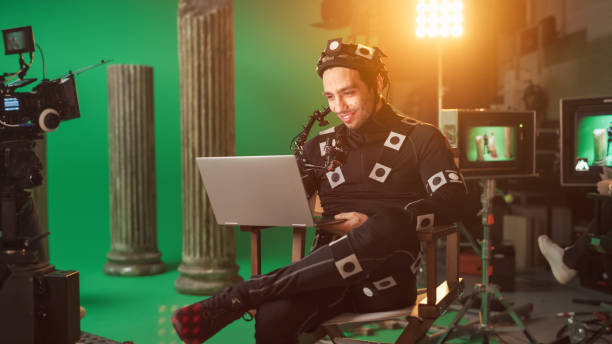 istock Handsome Smiling Actor Wearing Motion Capture Suit and Head Rig having Lunch Break, Sitting on Chair, Use Laptop. Studio High Budget Movie. On Film Studio Period Costume Drama Film Set 1265157480