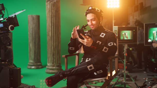 istock Handsome Smiling Actor Wearing Motion Capture Suit and Head Rig having Lunch Break, Sitting on Chair, Uses Smartphone. Studio High Budget Movie. On Film Studio Period Costume Drama Film Set 1265157482