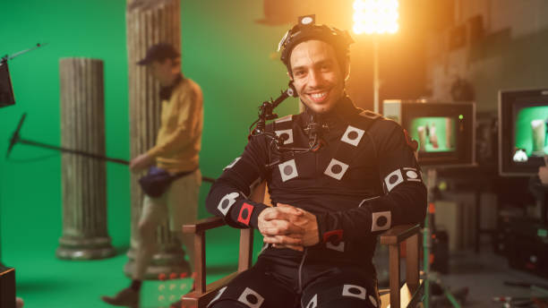 istock Handsome Smiling Actor Wearing Motion Capture Suit and Head Rig having Lunch Break, Sitting on Chair, Looking at Camera. Studio High Budget Movie. On Film Studio Period Costume Drama Film Set 1265157487