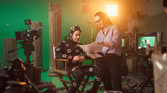 On Film Set: Prominent Female Director Explains Scene to Male Actor Wearing Motion Capture Suit and Playing Green Screen Scene in Superhero Movie. On Big Film Studio Professional Crew