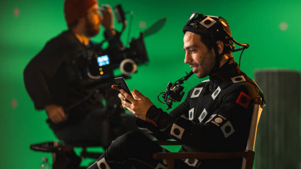 istock Handsome Smiling Actor Wearing Motion Capture Suit and Head Rig having Lunch Break, Sitting on Chair, Uses Smartphone. Studio High Budget Movie. On Film Studio Period Costume Drama Film Set 1265157532