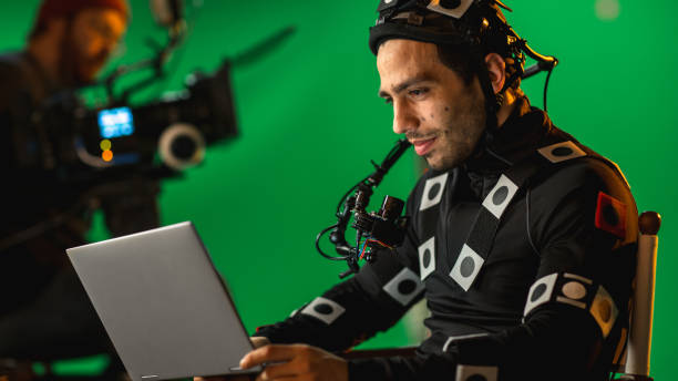 istock Handsome Smiling Actor Wearing Motion Capture Suit and Head Rig having Lunch Break, Sitting on Chair, Use Laptop. Studio High Budget Movie. On Film Studio Period Costume Drama Film Set 1265157564
