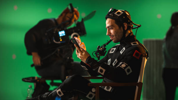 Handsome Smiling Actor Wearing Motion Capture Suit and Head Rig having Lunch Break, Sitting on Chair, Uses Smartphone. Studio High Budget Movie. On Film Studio Period Costume Drama Film Set