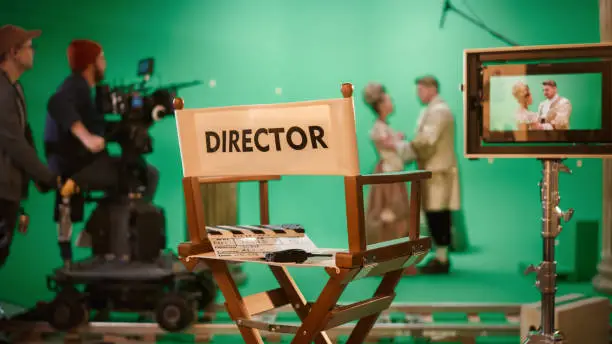 Photo of On Film Studio Set Focus on Empty Director's Chair. In the Background Professional Crew Shooting Historic Movie, Cameraman on Railway Trolley Shooting Green Screen Scene with Actors for History Movie