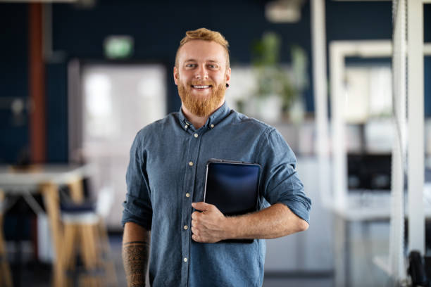 Portrait of a confident young businessman Portrait of a businessman with beard standing in office holding digital tablet. Confident male business executive in office looking at camera. looking at camera stock pictures, royalty-free photos & images