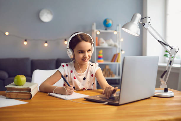 Home online schooling. Hardworking little girl in headset watching online lesson on laptop Home schooling concept. Hardworking little girl in headset watching online lesson on laptop, taking notes into her copybook indoors home schooling homework computer learning stock pictures, royalty-free photos & images