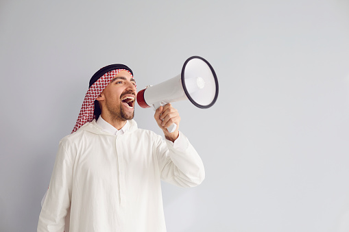 Hombre árabe con un megáfono en la mano gritando llamadas anuncia ofertas sobre un fondo gris para texto. photo