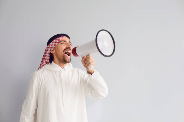 Arab man with a megaphone in hand shouting calls advertises offers on a gray background for text. Arab man with a megaphone in hand shouting calls advertises offers on a gray background for text. kaffiyeh stock pictures, royalty-free photos & images
