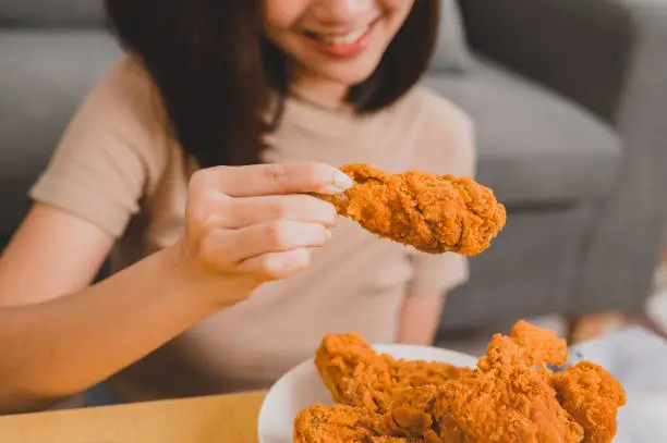 Photo of Eating fried chicken when takeout and delivery. Fast food takeaway back home.  Asian woman lifestyle in living room. Social distancing and new normal.