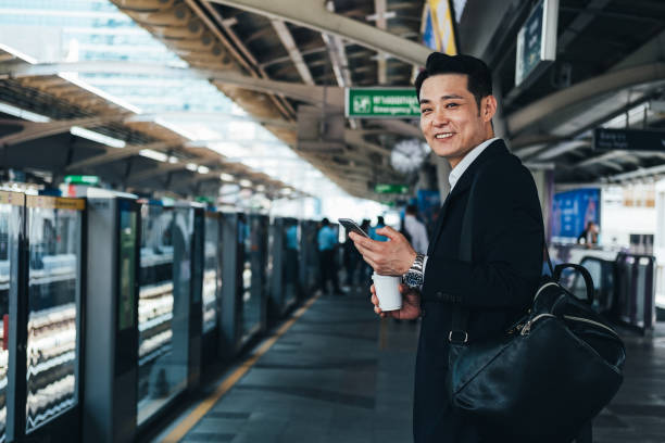 uśmiechnięty biznesmen trzymający jednorazową filiżankę kawy i telefon na dworcu kolejowym zdjęcie stockowe - bangkok mass transit system zdjęcia i obrazy z banku zdjęć