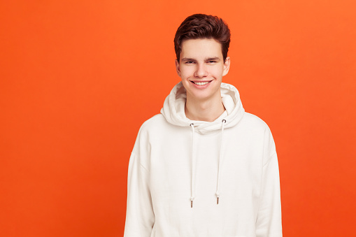 Smiling teenager in hoody looking at camera, positive and successful male person. Happy youth. Indoor studio shot isolated on orange background