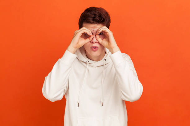 istock Funny curious young man in casual white sweatshirt looking far away through binoculars gesture, spying with open mouth, shocking content 1265192014