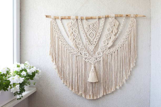 istock Handmade macrame. 100% cotton wall decoration with wooden stick hanging on a white wall.   Macrame braiding and cotton threads.  Female hobby.  ECO friendly modern knitting DIY natural decoration concept in the interior 1265192411