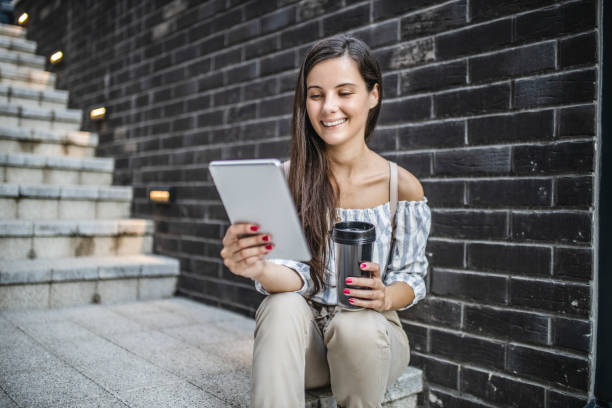 joven mujer hermosa sentada en las escaleras, usando tableta digital, bebiendo café, sonriendo y tomando un descanso - reading newspaper break tea fotografías e imágenes de stock