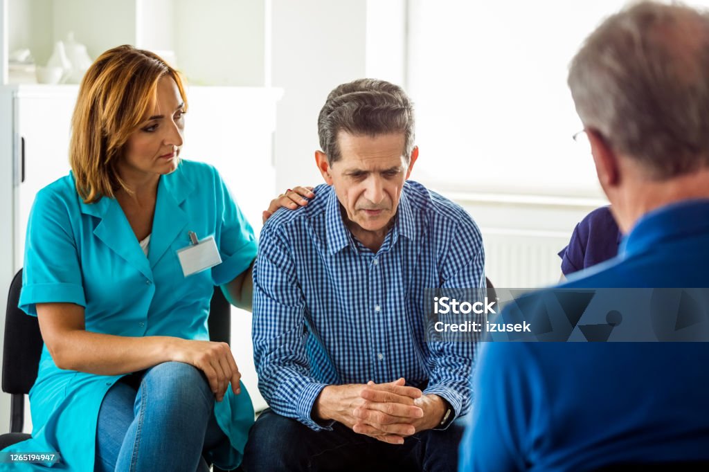 Senior man sharing issues during meeting Senior man sharing his issues with patients and nurse during meeting in nursing home. They are discussing about mental wellbeing. Nurse consoling an elderly man. Group Therapy Stock Photo