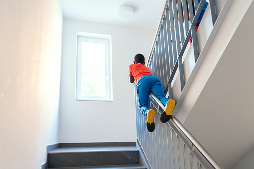 rear view on little boy with orange sports shirt slipping down balustrade in bright staircase