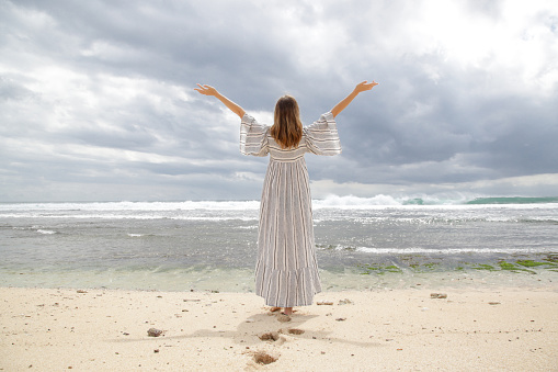 Woman standing on the beach with raised arms and looking at the horizon. Back view of woman in long white dress on the shore during gloomy summer day. Concept of freedom, happiness and positivity.