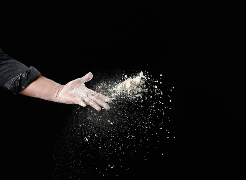 baker's male hand throws up a handful of white wheat flour on a black background, particles fly in different directions