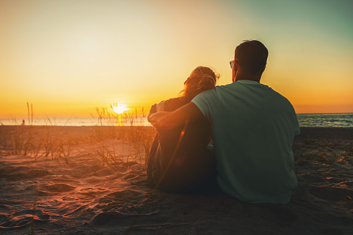 young lovers couple sitting in sand on beach at romantic golden sunset. back view