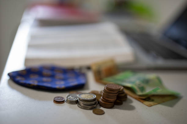 A close up of a office desk. On the desk is an open Bible, laptop, South African paper money and coins and a Covid 19 protective mask.