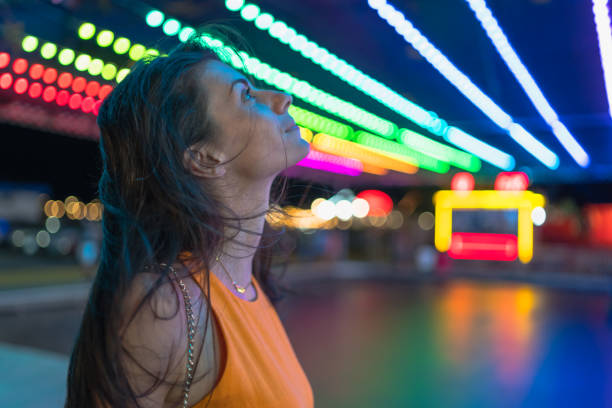 verticale de jeune femme joyeuse heureuse observant les lumières de carnaval. fond de lumières brouillées colorées - ferris wheel flash photos et images de collection