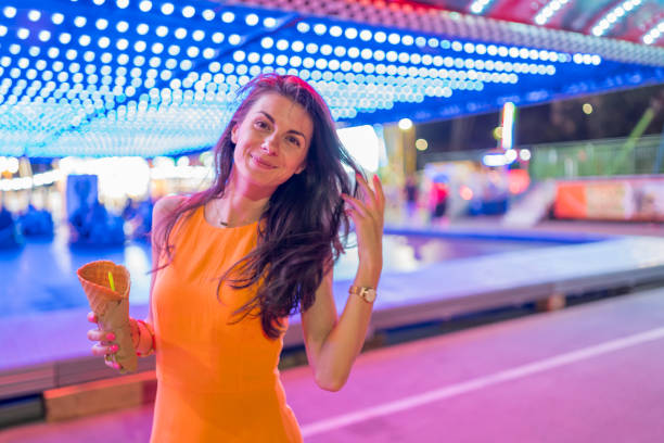verticale de jeune femme joyeuse rectifiée avec la robe orange lumineuse posant devant des voitures de pare-chocs au parc d’attraction, retenant la crème glacée - ferris wheel flash photos et images de collection