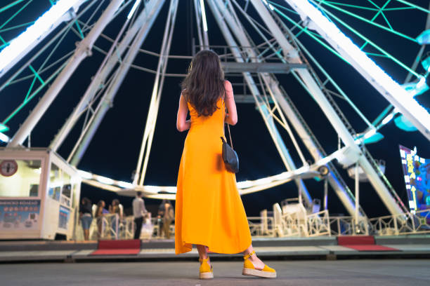 vue arrière de la femme en forme rectifiée avec la robe orange, restant devant la grande roue. plan de nuit du parc d’attractions illuminé - ferris wheel flash photos et images de collection