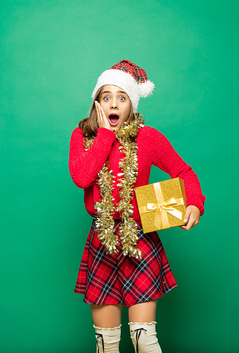 Excited teenager wearing funny christmas clothes, holding gift and looking at camera with mouth open. Studio shot against green background.