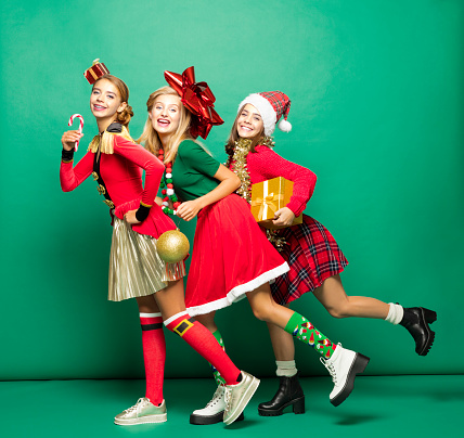 Three teenagers wearing funny Christmas clothes, holding presents and bauble laughing at the camera. Studio shot against green background.
