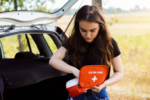 Young attractive girl stands near car with open back door and emergency lights is on, tries to find something in first aid kit Concept of car crash and giving first aid first aid photos stock pictures, royalty-free photos & images