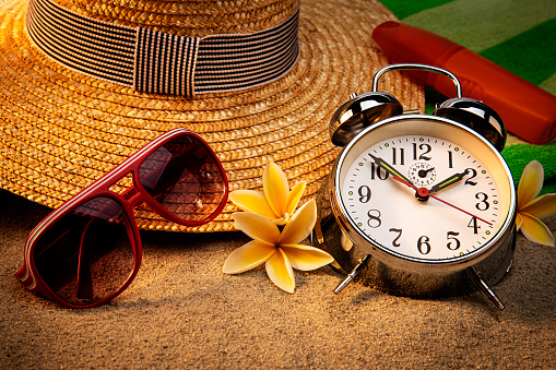 This is a photograph of an alarm clock, sandals, sunglasses, a hat and a palm branch on a sandy beach