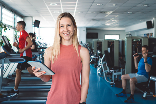Attractive personal coach in an indoor gym, group people in the background, training, using fitness equipment. Professional fitness instructor. Helping people get fit and exercise well is my professional occupation. Portrait photo. Using modern technology in workout plans.