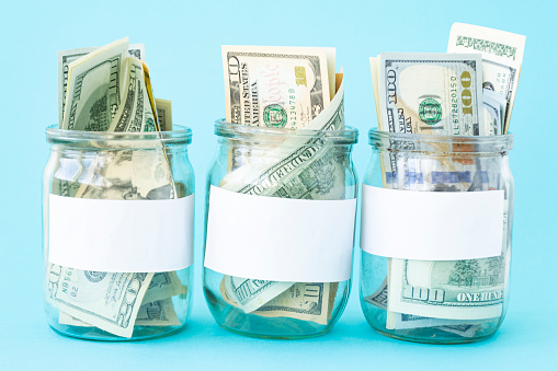 Three Piggy Bank from a Glass Jar for Life. New dollars sticking out of piggy banks on a blue background. The banks have white sheets for inscriptions. Mockup. Copy space. Economy concept.