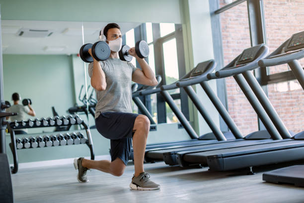 atleta masculino usando mascarilla facial mientras hace ejercicio con pesas de mano en posición de estocada en un gimnasio. - body building exercises audio fotografías e imágenes de stock