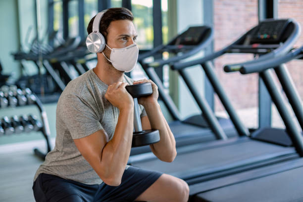 istock Athletic man wearing face mask while using dumbbell and exercising squats in a gym. 1265281091