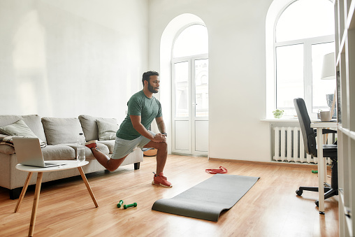 Get Fit Online. Full length shot of young active man watching online video training on laptop, exercising during morning workout at home. Sport, healthy lifestyle. Horizontal shot